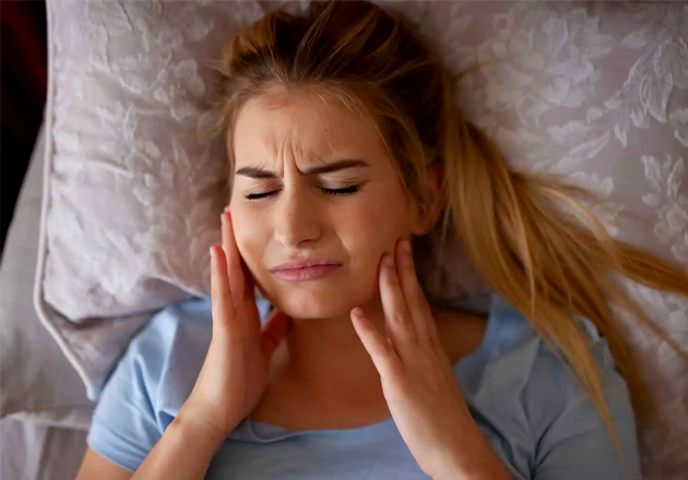 Woman lying on pillow experiencing jaw pain, holding cheeks with both hands in discomfort.