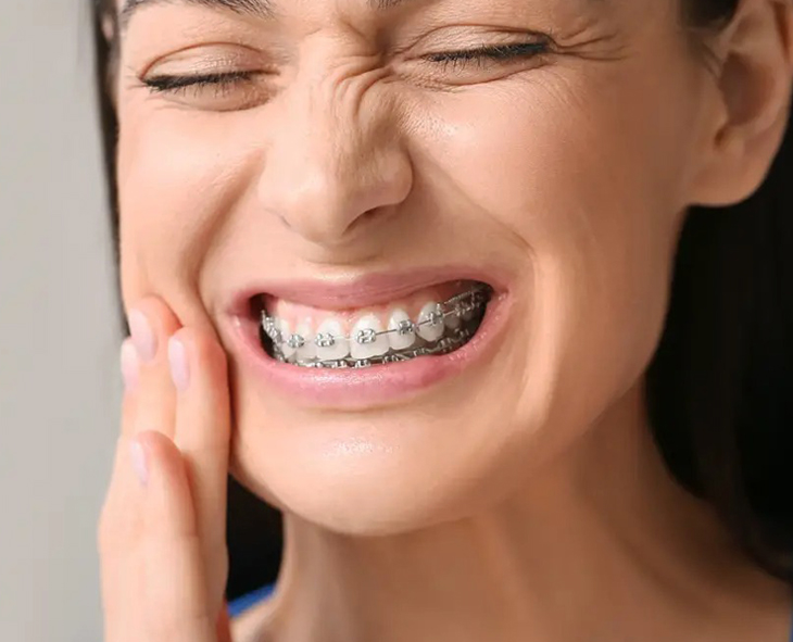 Woman with braces touching her cheek, wincing in dental pain or discomfort indoors.