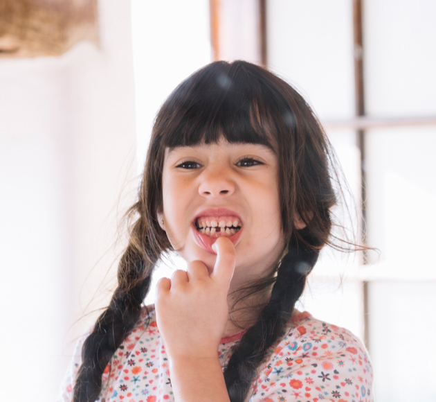 Young girl with dark hair smiling with finger in mouth, casual indoor setting.