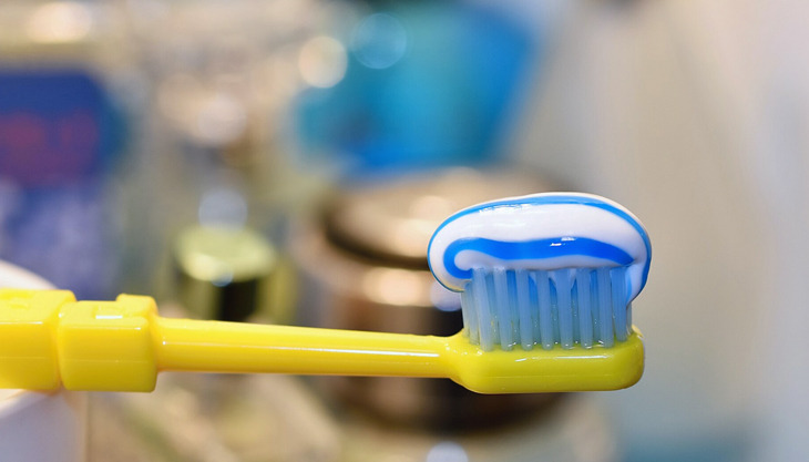 Yellow toothbrush with blue toothpaste against blurred bathroom background.
