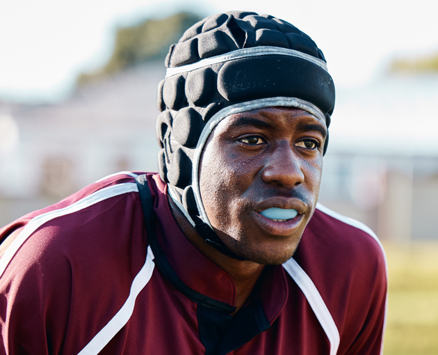 Male athlete wearing maroon jersey and protective headgear with mouthguard, outdoor sports field setting.