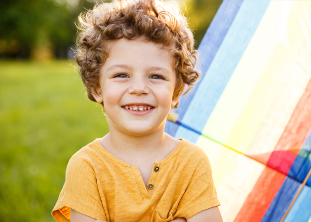 Happy child in yellow shirt smiling outdoors with colorful kite in park setting.