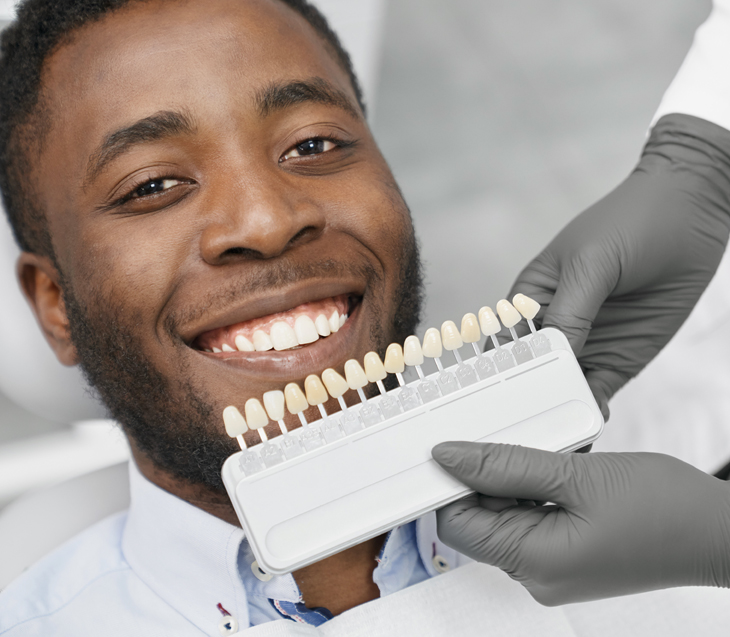 Dentist holding tooth shade guide against smiling patient's teeth during consultation.