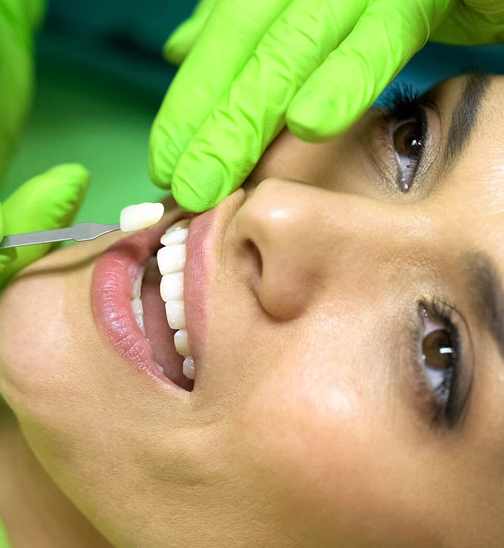Person receiving dental examination with gloved hands holding mouth open against green background.