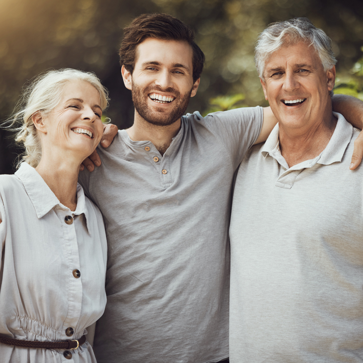 Three generations of a family smiling together outdoors in natural sunlight.