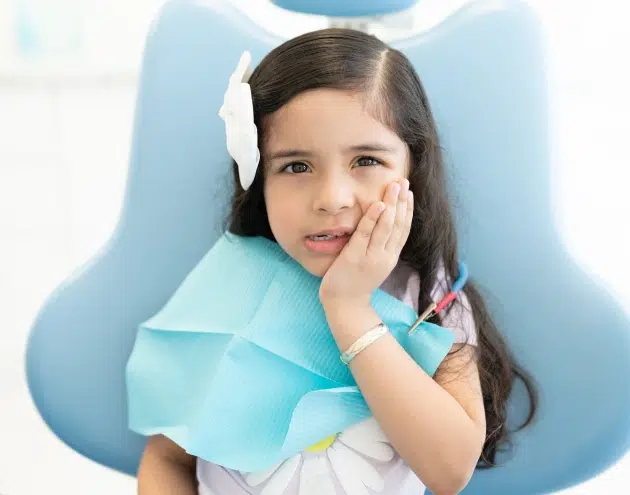 Young girl smiling at dental office, hand on cheek, wearing blue bib in chair.